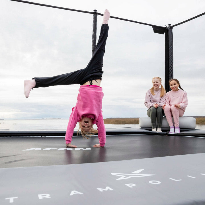 Zwei Mädchen sitzen auf dem aufblasbaren Acon X Triangle Block. Das Mädchen steht mit den Händen auf dem Trampolin.